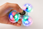 A hand holding a three-spoke LED fidget spinner with glowing multicolor lights on a plain white background.