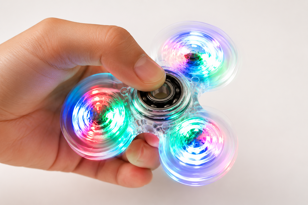 A hand holding a three-spoke LED fidget spinner with glowing multicolor lights on a plain white background.