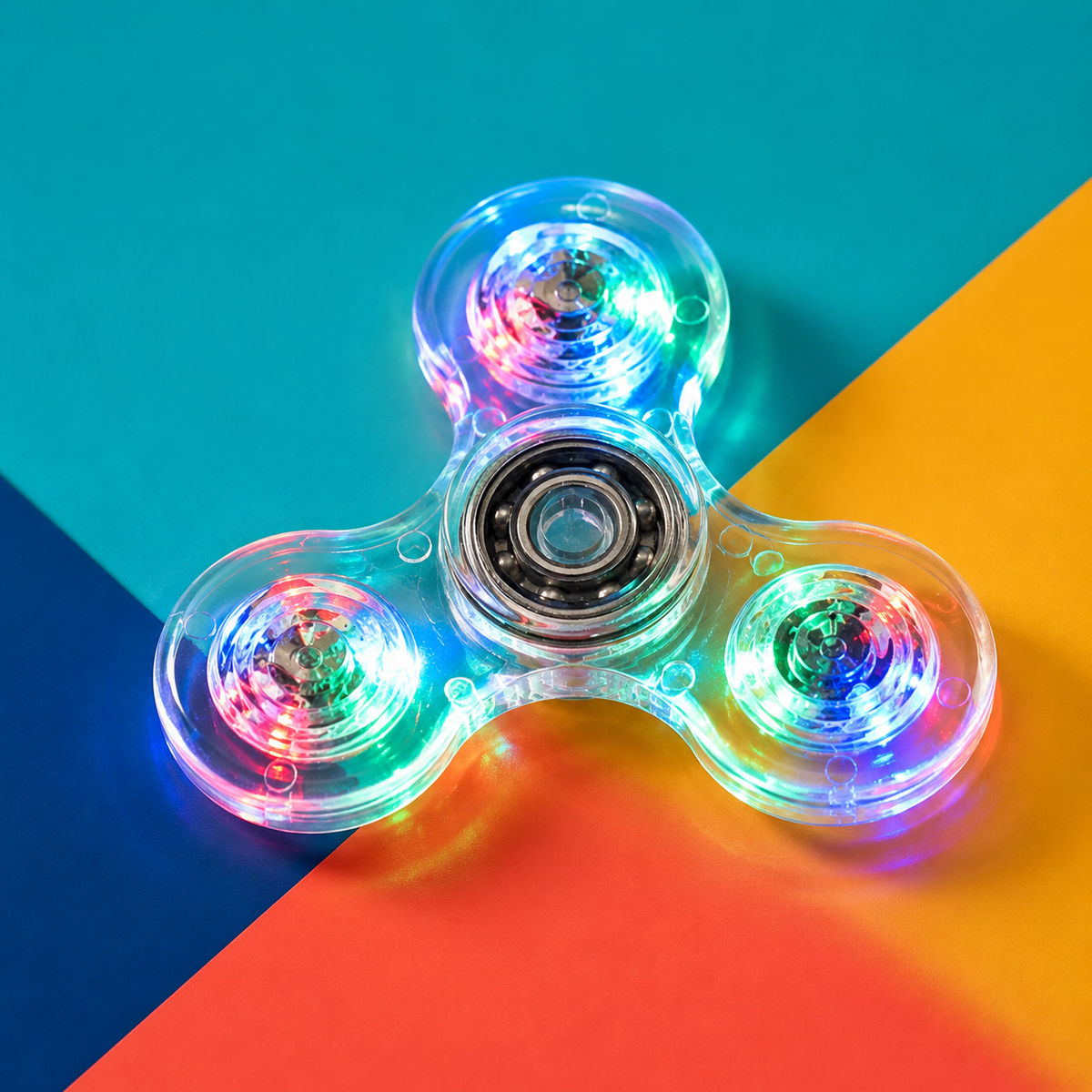 A three-spoke transparent fidget spinner with glowing multicolor LED lights, centered on a diagonally split teal, orange, and dark blue background.