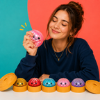 A woman in a navy sweatshirt holds a pink glittery, kawaii-style squishy toy while five more colorful squishies in orange holders sit lined up on a white table against a bright teal and coral background.